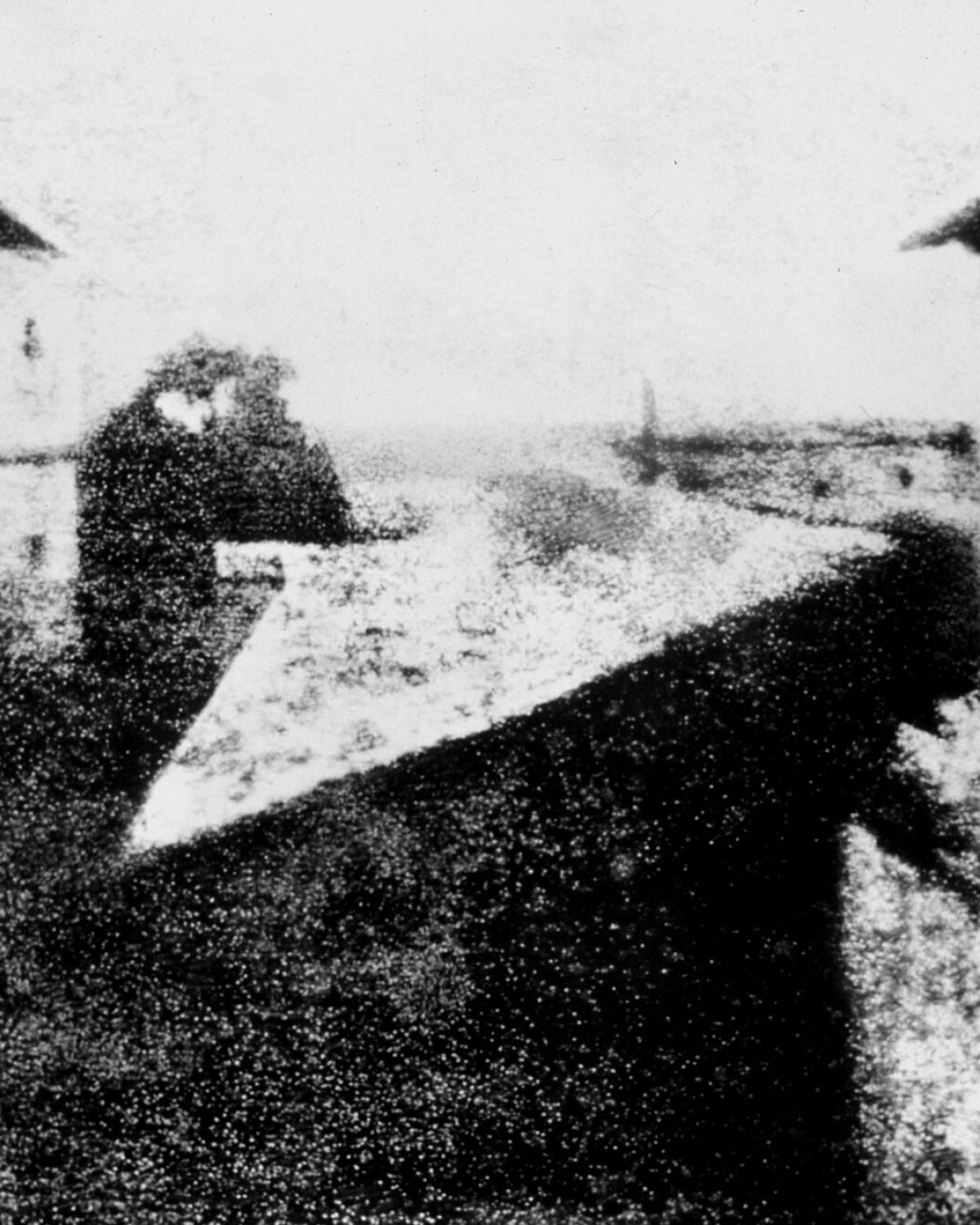 Vue floue et granuleuse, en noir et blanc, des toits et des bâtiments d’une propriété, prise depuis une fenêtre en hauteur.